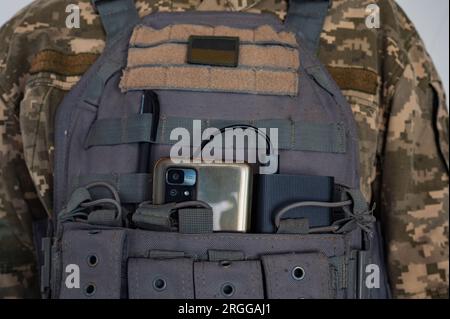 Ukrainischer Soldat in grauer kugelsicherer Jacke mit Banner der ukrainischen Flagge, Smartphone und Powerbank. Stockfoto