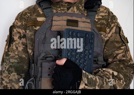 Soldat in grauer kugelsicherer Jacke mit Icon der ukrainischen Flagge und Tastatur im Arm in schwarzem Handschuh. Stockfoto