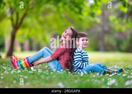 Fröhliche Kinder Junge und Mädchen in Gummistiefeln spielen draußen im grünen Park mit blühenden Blüten Stockfoto
