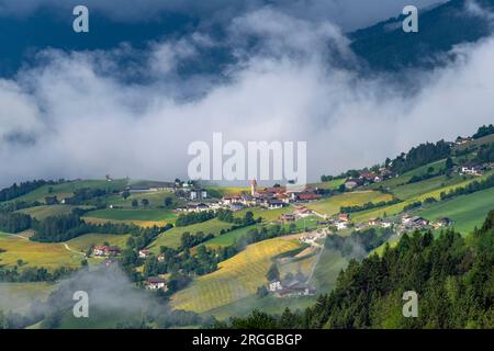 Panoramablick auf das Dorf Aicha oder Aica in Südtirol, Italien, mit charakteristischem Kirchturm umgeben von Weiden und tief hängenden weißen Wolken Stockfoto