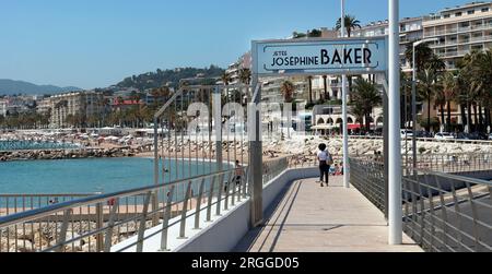 Eine Frau schlendert entlang des Josephine Baker Jetty in Cannes. Der öffentliche Strand von Cannes Old Port ist links eingefasst; Hotels mit Blick auf die Bucht von Cannes Hintergrund Stockfoto