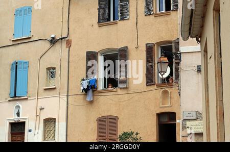 Ein kleines Mädchen schaut aus einem verschlossenen Fenster; Waschen hängt am Verschluss in der Altstadt von Fréjus, einem provenzalischen Dorf im Var, Côte d'Azur Stockfoto