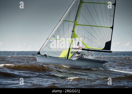 Der Blick auf das Sportsegelboot ist bei Sonnenuntergang hoch, Sportler neigen das Boot, Geschwindigkeit und Spritzwasser, Reflexion der Sonne auf der Wasseroberfläche Stockfoto