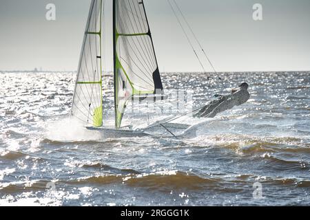 Der Blick auf das Sportsegelboot ist bei Sonnenuntergang hoch, Sportler neigen das Boot, Geschwindigkeit und Spritzwasser, Reflexion der Sonne auf der Wasseroberfläche Stockfoto