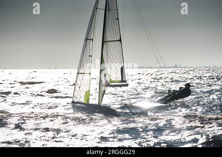 Der Blick auf das Sportsegelboot ist bei Sonnenuntergang hoch, Sportler neigen das Boot, Geschwindigkeit und Spritzwasser, Reflexion der Sonne auf der Wasseroberfläche Stockfoto