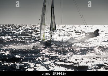 Der Blick auf das Sportsegelboot ist bei Sonnenuntergang hoch, Sportler neigen das Boot, Geschwindigkeit und Spritzwasser, Reflexion der Sonne auf der Wasseroberfläche Stockfoto