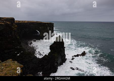 Lange Exposition von den Klippen in Island mit einem orangefarbenen Leuchtturm am Horizont. Stockfoto