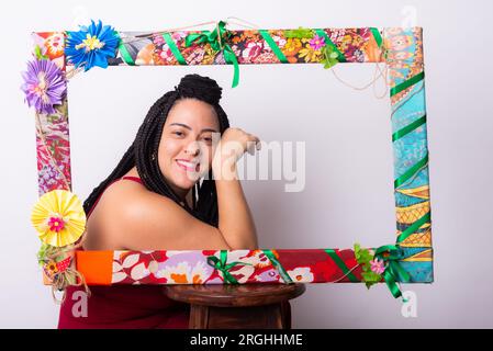 Foto einer wunderschönen Frau mit Zöpfen im Haar hinter einem Rahmen, der mit Blumen und farbenfrohen Tüchern in horizontaler Position dekoriert ist. Weiße Backe Stockfoto