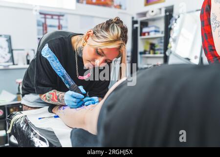 Erwachsene, professionelle Tätowierungskünstlerin, die das Bein einer jungen Frau tätowiert. Fokussierte Künstlerin mit einer Tattooing-Waffe. Hochwertiges Foto Stockfoto