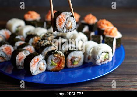 Schöne weibliche Hände nehmen Sushi mit Essstäbchen. Dunkler Hintergrund. Holzplanke. Seitenansicht. Horizontale Ansicht Blaues Schild. Sushi. Verschiedene Stockfoto