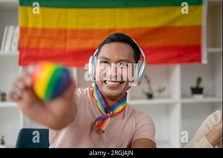 Nahaufnahme eines attraktiven und glücklichen asiatischen Schwulen mit Kopfhörern, der das LGBT-Herz in seinem Zimmer hält und an der Wand die LGBT-Flagge trägt. Vielfalt, ho Stockfoto