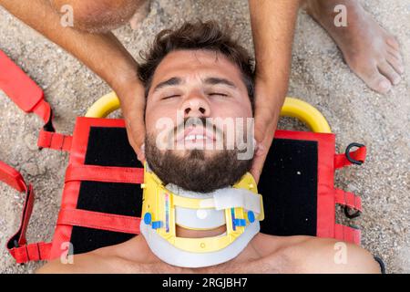 Draufsicht auf einen Mann mit einem verletzten medizinischen Halsband, der am Sandstrand liegt Stockfoto