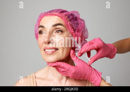 Porträt einer Frau mittleren Alters mit rosa Kappen für kosmetische Behandlungen bei Schönheitsinjektionen. Hände in rosa Handschuhen halten eine Spritze. Gesichtstraining. Stockfoto