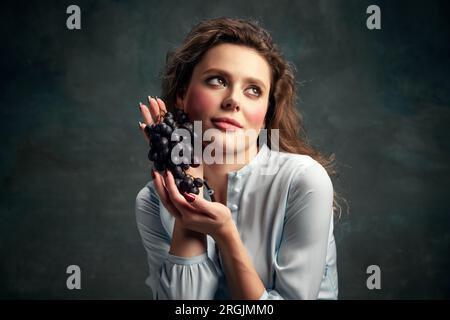Wunderschöne, glamouröse junge Frau in einem blauen historischen Kleid, die Trauben präsentiert und wegschaut. Essensstimmung. ERAS-Vergleich Stockfoto