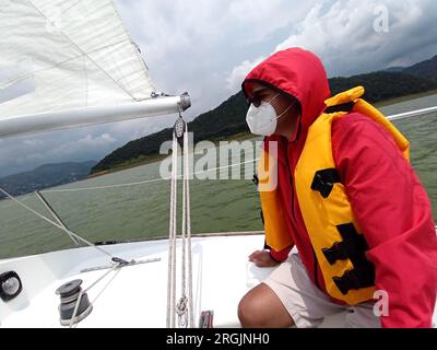 Ein erwachsener Latino-Mann fährt ein Segelboot auf dem See mit Windjacke und Schwimmweste, trägt Gesichtsmasken in der neuen Normalität aufgrund der Covid-19-Pandemie Stockfoto