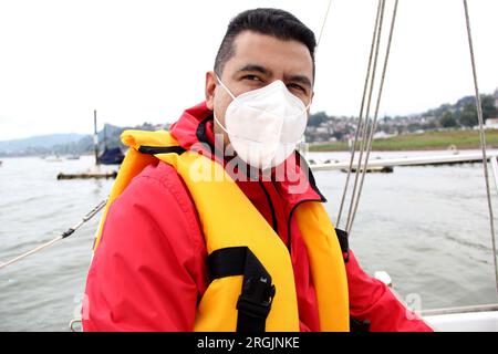 Ein erwachsener Latino-Mann fährt ein Segelboot auf dem See mit Windjacke und Schwimmweste, trägt Gesichtsmasken in der neuen Normalität aufgrund der Covid-19-Pandemie Stockfoto