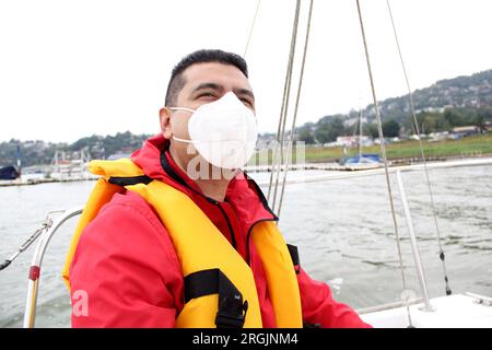 Ein erwachsener Latino-Mann fährt ein Segelboot auf dem See mit Windjacke und Schwimmweste, trägt Gesichtsmasken in der neuen Normalität aufgrund der Covid-19-Pandemie Stockfoto