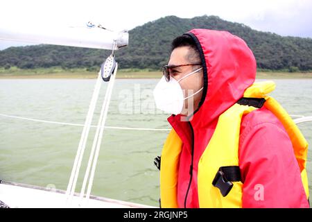 Ein erwachsener Latino-Mann fährt ein Segelboot auf dem See mit Windjacke und Schwimmweste, trägt Gesichtsmasken in der neuen Normalität aufgrund der Covid-19-Pandemie Stockfoto