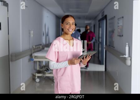 Porträt einer glücklichen, birassischen Ärztin, die Kittel trägt und Tabletten im Flur des Krankenhauses hält Stockfoto