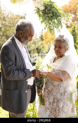 Ein glücklicher älterer Bräutigam, der bei einer sonnigen Hochzeitszeremonie im Freien den Ring auf den Finger der Braut legt Stockfoto