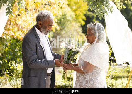 Glückliche ältere birassische Braut und Bräutigam während der Hochzeitszeremonie im sonnigen Garten Stockfoto