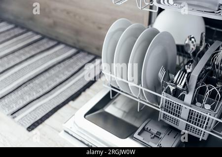 Vervollständigung der geöffneten automatischen Edelstahl eingebauten Geschirrspülmaschine in der modernen Küche mit sauberem Utensil. Weiße Teller, Geschirr, Becher, Metallgabeln und Messer. Haushalt, Haushalt, Hausarbeit Stockfoto