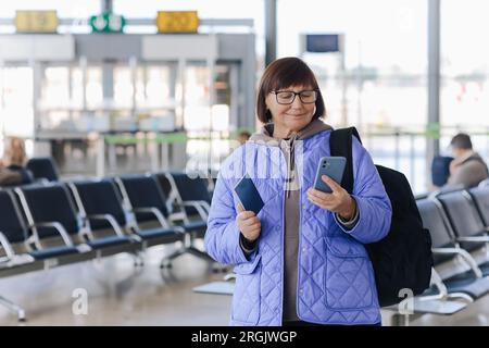 Reife Reisende mit Handgepäck, Rucksack und Reisepass checken online die Abflugzeit oder checken am Flughafen per Smartphone ein Stockfoto