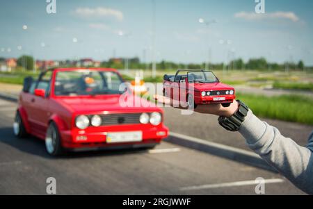 Die Hand auf der linken Seite hält ein rotes Volkswagen Golf MK2 Cabriolet und ein großes echtes Auto, das im Hintergrund steht. Stockfoto