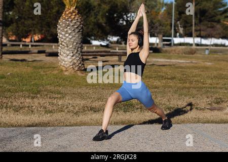 Eine junge gesunde Frau in Sportbekleidung hört Musik über Kopfhörer, während sie im Park mit Palmen Fitness-Squat-Beinübungen im Freien macht. Workout für st Stockfoto