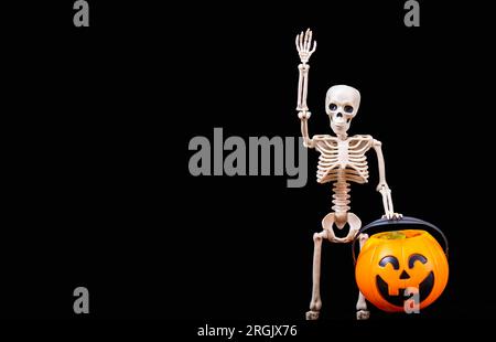 Ein menschliches Skelett, das winkt, einen orangefarbenen Halloween-Kürbis auf schwarzem Hintergrund hält. Halloween-Grußkarte Stockfoto