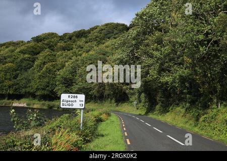 Landschaft mit einer Straße, die von einem See und Bäumen in Laub begrenzt wird, mit zweisprachigen Straßenschildern, die im ländlichen County Leitrim, Irland, zu sehen sind Stockfoto