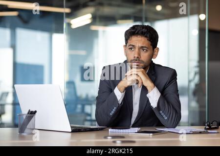 Ernsthaft denkender arabischer Geschäftsmann am Arbeitsplatz, Mann sitzt am Schreibtisch mit Laptop im Business-Anzug und konzentriert sich auf die Lösung finanzieller Probleme. Stockfoto