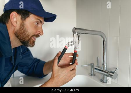 Professioneller Klempner in blauer Uniform, der kaputte Wasserhähne in der Küche fixiert. Reparaturservicekonzept Stockfoto