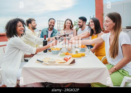 Fröhliche multikulturelle Gruppe junger erwachsener Freunde, die zusammen eine Dinnerparty mit gesundem Essen und Rotweingläsern feiern. Freunde, die beim Geburtstagsessen Spaß und Jubel haben. Hochwertiges Foto Stockfoto