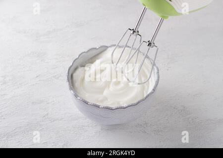 Eine hellblaue Schüssel mit ausgetretenem Eiweiß mit weichen Spitzen und einem Mixer auf hellgrauem Hintergrund. Bühne der Zubereitung köstlicher hausgemachter Desserts Stockfoto