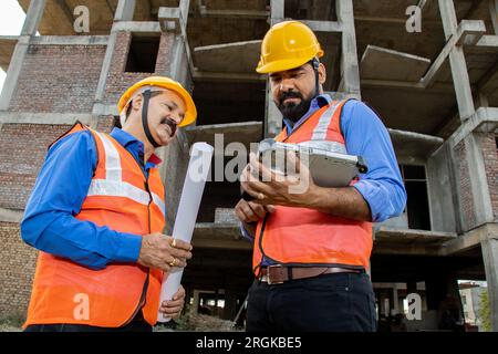 Zwei männliche Bauingenieure oder Architekten mit Helm und Weste mit einem digitalen Tablet-Blueprint auf der Baustelle, die über Immobilienprojekte diskutieren Stockfoto