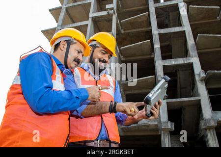 Zwei männliche Bauingenieure oder Architekten mit Helm und Weste mit einem digitalen Tablet-Blueprint auf der Baustelle, die über Immobilienprojekte diskutieren Stockfoto