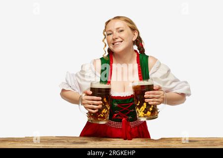 Freundliche junge Frau, die volkstümliches Dirndl mit zwei Biertassen auf weißem Hintergrund trägt. Feiertagsveranstaltung, Oktoberfest Stockfoto