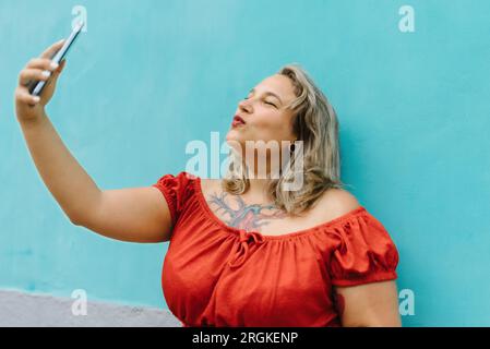 Seitenansicht einer jungen Frau in Übergröße in legerer Kleidung, die Selfie im runden Mund macht und mit dem Smartphone posiert, während sie draußen auf blauem Wandhintergrund steht Stockfoto
