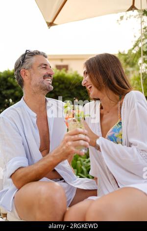 Fröhliches, ethnisch-romantisches Paar im Sommer, das erfrischende Cocktails trinkt und während der Feiertage auf den Liegen lacht Stockfoto