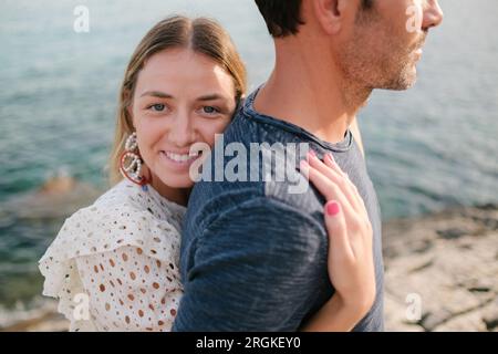 Junge Frau lächelt und schaut in die Kamera, während sie im Sommer den Mann am Meer umarmt Stockfoto