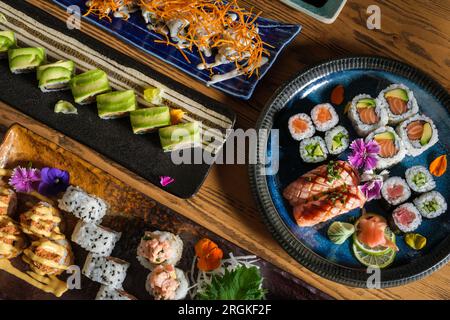 Blick von oben auf köstliche Sushi-Brötchen mit Fisch und verschiedenen Füllungen, serviert auf einem Tablett mit dekorativen Blumen und Wasabi mit Ingwer Stockfoto