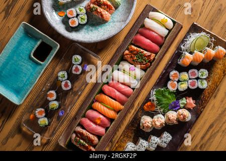 Blick von oben auf Sushi und Brötchen mit verschiedenen frischen Fischen, serviert auf Tabletts auf einem Holztisch mit Sojasoße im Café Stockfoto