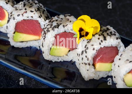 Leckere Sushi-Brötchen mit Avocado und Thunfisch, serviert auf einem blauen Tablett mit Blumen auf dem Tisch im Café vor schwarzem Hintergrund Stockfoto