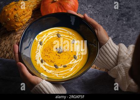 Hohes Erntegut, nicht wiedererkennbares Weibchen, mit einer Schüssel leckerer OrangenKürbissuppe mit Samen am dunklen Tisch Stockfoto