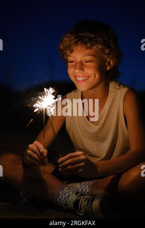 Fröhlicher Junge, der nachts mit glühenden Glitzern sitzt und festliche Anlässe feiert Stockfoto