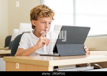 Intelligentes Schulkind mit Stift auf dem Tablet, während es in einem modernen Klassenzimmer in der Schule studiert Stockfoto
