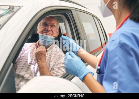 Schneiden Sie nicht erkennbare weibliche Sanitäter mit Wattestäbchen im Gespräch mit älteren männlichen Fahrer vor der Analyse für COVID 19 im Auto Stockfoto
