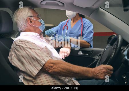 Anonyme weibliche Medikerin in Atemschutzmaske mit Spritze zur Impfung älterer männlicher Patienten im Auto während der COVID 19-Pandemie ausschneiden Stockfoto