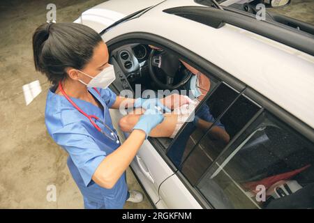 Von oben von weiblichem Sanitäter in Uniform mit Spritze iminokulierenden männlichen Patienten in Auto während Coronavirus Pandemie Stockfoto